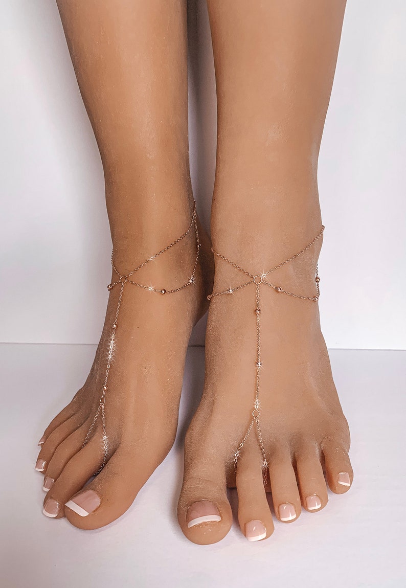Peut inclure: Gros plan sur des pieds orn&eacute;s de sandales dor&eacute;es. Les sandales sont compos&eacute;es d'une cha&icirc;ne avec de petites perles et des d&eacute;tails en forme d'&eacute;toiles, reliant la cheville et s'&eacute;tendant jusqu'aux orteils. Les ongles sont peints avec une manucure fran&ccedil;aise.