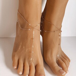 Peut inclure: Bijoux de pied dor&eacute;s d&eacute;licats. Les bracelets de cheville comportent plusieurs brins avec de petits ornements scintillants et une cha&icirc;ne pendante qui se connecte &agrave; l'orteil. Les bijoux sont port&eacute;s sur des pieds avec des ongles d'orteils manucur&eacute;s.