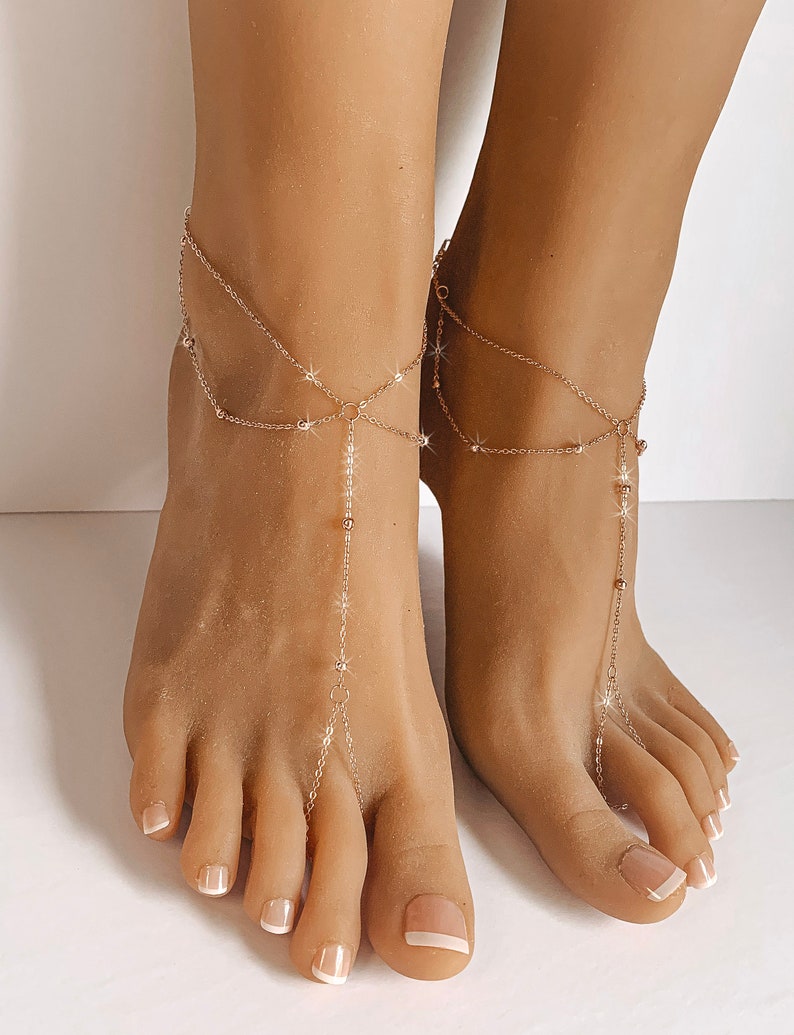 Peut inclure: Bijoux de pied dor&eacute;s d&eacute;licats. Les bracelets de cheville comportent plusieurs brins avec de petits ornements scintillants et une cha&icirc;ne pendante qui se connecte &agrave; l'orteil. Les bijoux sont port&eacute;s sur des pieds avec des ongles d'orteils manucur&eacute;s.