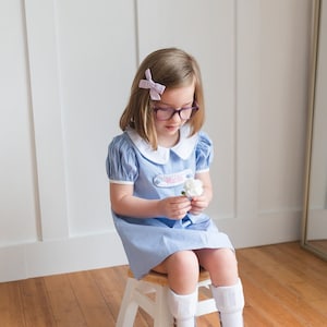 May include: A young girl wearing a light blue dress with a white collar and sleeves, seated on a small wooden stool. The dress has a name tag that says "Matilda". She wears glasses, a bow, white socks, and white sneakers. She holds a white flower.