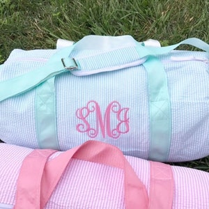 May include: Two striped canvas duffel bags, one in light blue with a pink monogram "SMC" and the other in pink. Both bags have a single zipper and a top handle.