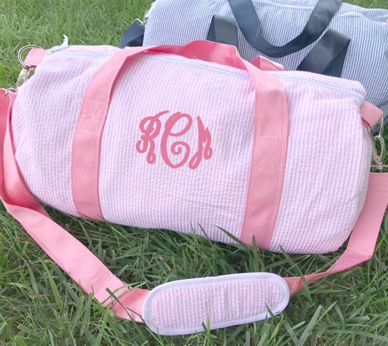 May include: Pink and white striped duffel bag with a pink monogram "KCM" on the front. The bag has a pink strap and a pink and white striped handle.