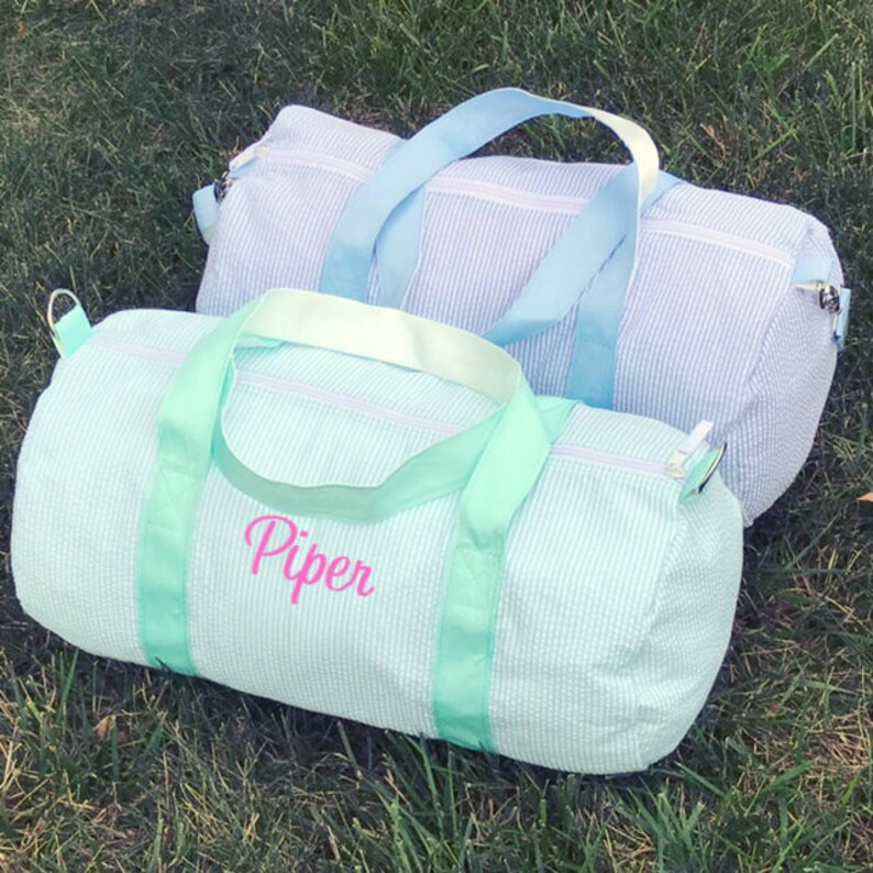 May include: Two striped canvas duffel bags. The bag on the bottom is light blue with a green stripe and the name "Piper" embroidered on the front. The bag on top is light blue with a light blue stripe.