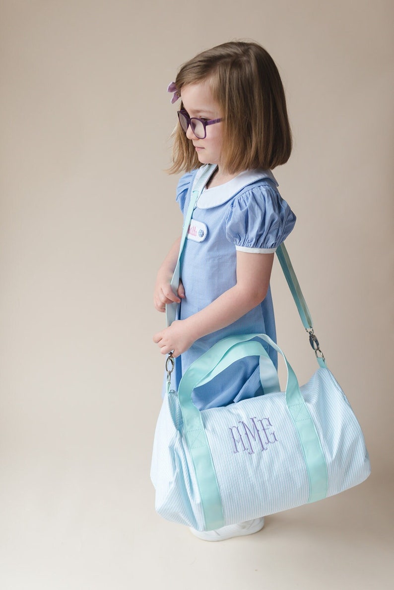 May include: A light blue and white striped duffel bag with a light blue strap and a light blue handle. The bag has a purple monogram that says "EMMA".
