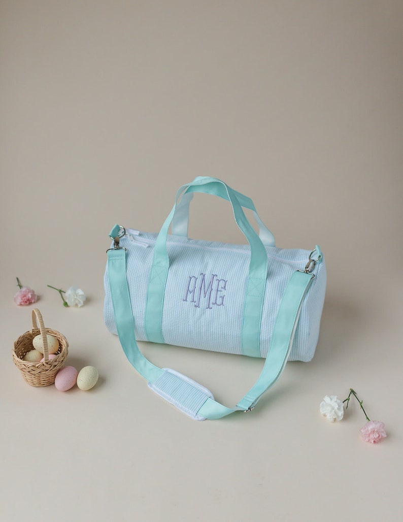 May include: A light blue striped duffel bag with a light blue strap and a monogram that says "AME" in purple thread.