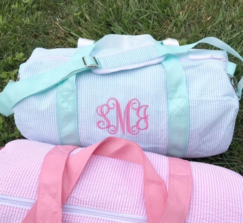 May include: Two striped canvas duffel bags, one in light blue with a pink monogram "SMC" and the other in pink. Both bags have a single zipper and a top handle.