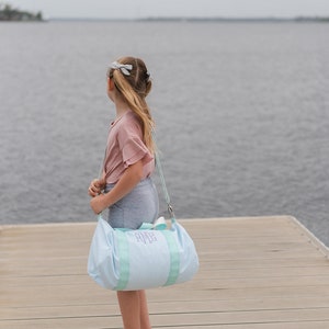 May include: A young person standing on a wooden dock, looking out at the water. They are carrying a light blue and white striped duffel bag with the monogram "AMG" embroidered on it.