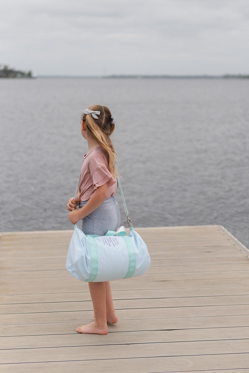 May include: A young person standing on a wooden dock, looking out at the water. They are carrying a light blue and white striped duffel bag with the monogram "AMG" embroidered on it.