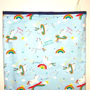 May include: A light blue fabric with a repeating pattern of white unicorns, rainbows, and stars. The unicorns are jumping over rainbows, surfing on rainbows, and holding hoops. The text "Hola time", "You're amazing", "Jump!" and "Surf the rainbow" is printed on the fabric.