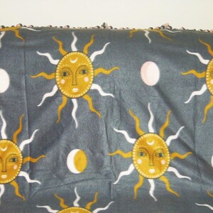 May include: A gray fleece blanket with a tie-dye fringe border. The blanket features a repeating pattern of yellow suns with white rays and pink moons.