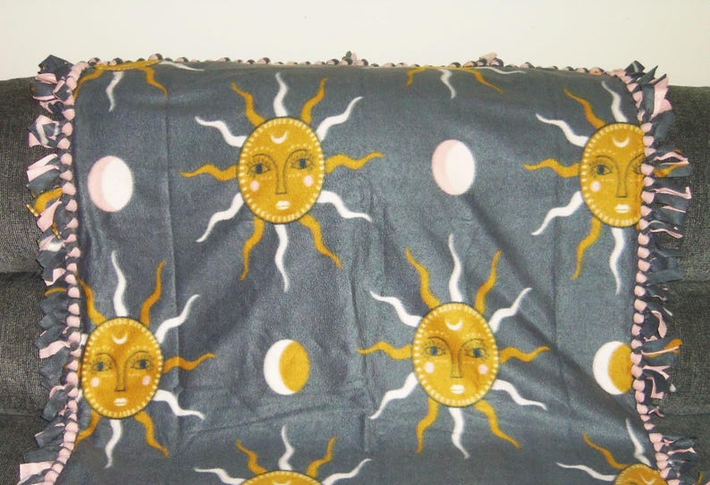 May include: A gray fleece blanket with a tie-dye fringe border. The blanket features a repeating pattern of yellow suns with white rays and pink moons.