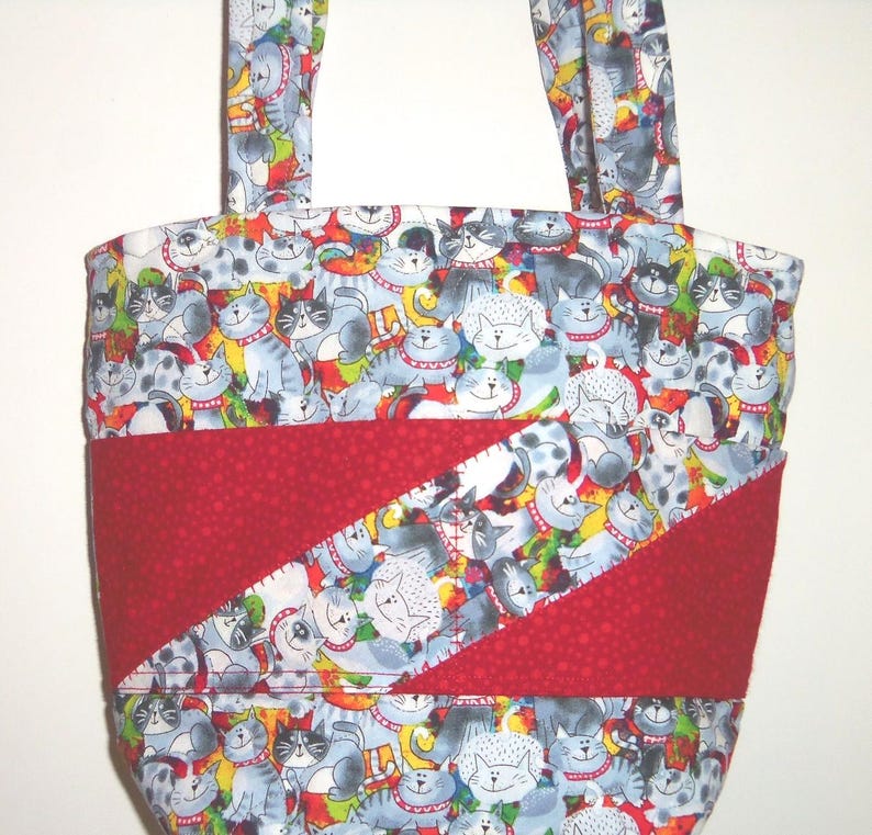 May include: A fabric tote bag featuring a repeating pattern of cartoon cats in shades of gray, white, and black with red collars. The bag includes a diagonal red fabric panel across the center with visible stitching and two matching fabric handles.