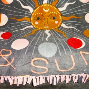 May include: A gray fleece blanket with a pink fringe border. The blanket features a sun and moon design with the words "& SUN" in the center.