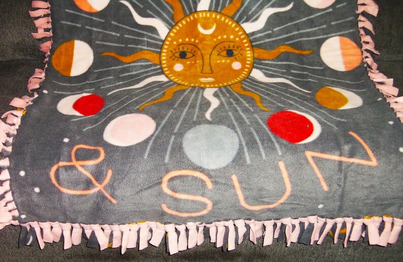 May include: A gray fleece blanket with a pink fringe border. The blanket features a sun and moon design with the words "& SUN" in the center.