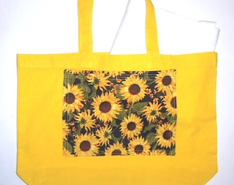 May include: A bright yellow tote bag featuring a rectangular front pocket with a dark background and a repeating sunflower pattern. The bag has two matching yellow shoulder straps and contains a white rectangular object inside.