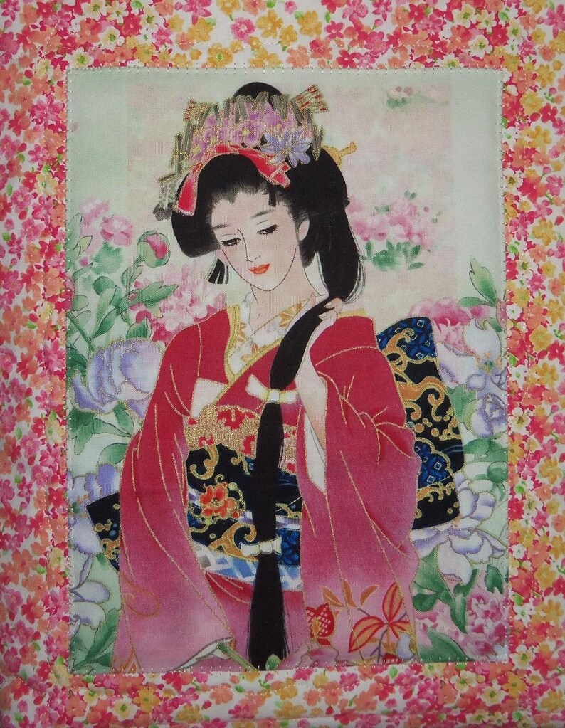 May include: A fabric wall hanging featuring a central illustration of a person with long black hair wearing a red and black kimono with gold accents. The figure is surrounded by pink and purple flowers, framed by a border of small floral patterns.