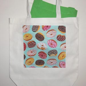 May include: A white tote bag featuring a rectangular patch with a light blue background and a repeating pattern of various donuts with colorful icing, sprinkles, and chocolate drizzles. A bright green fabric item is tucked inside the bag.