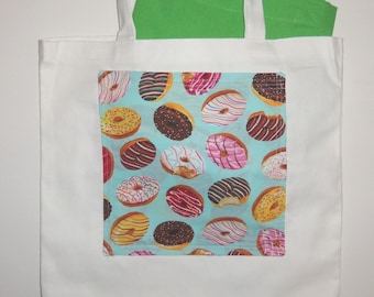 May include: A white tote bag featuring a rectangular patch with a light blue background and a repeating pattern of various doughnuts with colourful icing, sprinkles, and chocolate drizzles. A bright green fabric item is tucked inside the bag.