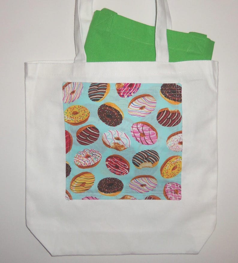 May include: A white tote bag featuring a rectangular patch with a light blue background and a repeating pattern of various donuts with colorful icing, sprinkles, and chocolate drizzles. A bright green fabric item is tucked inside the bag.