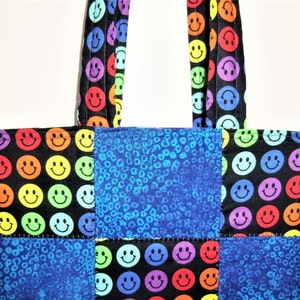 Puede incluir: Un bolso tote con un dise&ntilde;o de patchwork de tela negra con un patr&oacute;n de caras sonrientes multicolores y tela con estampado de leopardo azul. El bolso incluye dos asas de tela a juego con el mismo patr&oacute;n de caras sonrientes sobre fondo negro.