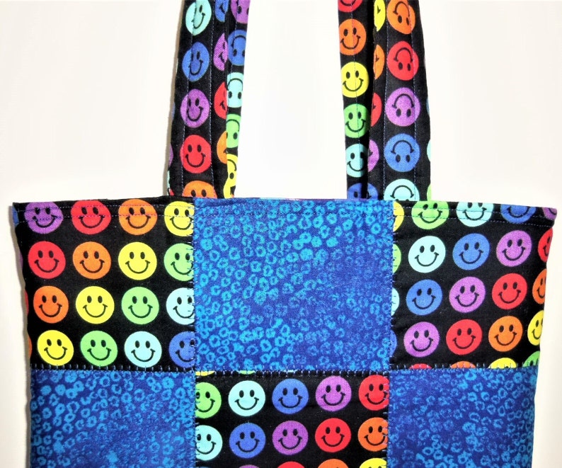 Puede incluir: Un bolso tote con un dise&ntilde;o de patchwork de tela negra con un patr&oacute;n de caras sonrientes multicolores y tela con estampado de leopardo azul. El bolso incluye dos asas de tela a juego con el mismo patr&oacute;n de caras sonrientes sobre fondo negro.