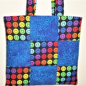 Puede incluir: Un bolso tote acolchado con un dise&ntilde;o de patchwork de tela negra estampada con caras sonrientes coloridas y tela azul con un patr&oacute;n animal texturizado. El bolso incluye dos asas de tela a juego en la parte superior para llevarlo.