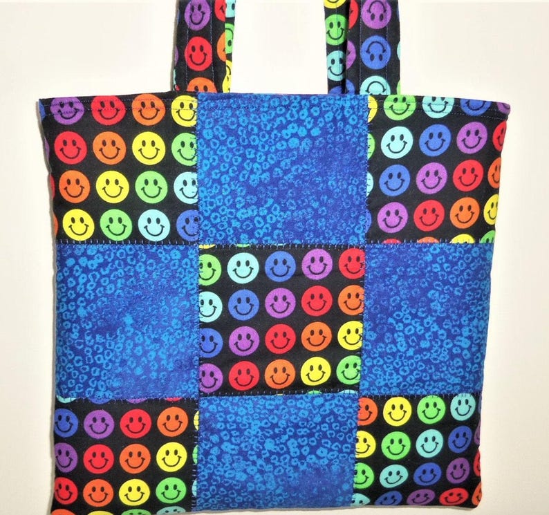 Puede incluir: Un bolso tote acolchado con un dise&ntilde;o de patchwork de tela negra estampada con caras sonrientes coloridas y tela azul con un patr&oacute;n animal texturizado. El bolso incluye dos asas de tela a juego en la parte superior para llevarlo.