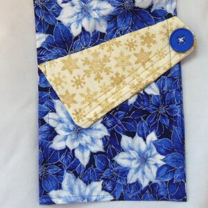 May include: A rectangular fabric towel featuring a vibrant blue and white poinsettia floral pattern. A folded top flap in cream fabric with a gold snowflake print is secured by a single, round, bright blue button on the right side.