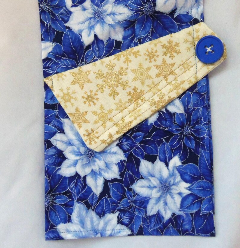 May include: A rectangular fabric towel featuring a vibrant blue and white poinsettia floral pattern. A folded top flap in cream fabric with a gold snowflake print is secured by a single, round, bright blue button on the right side.