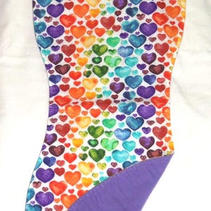 May include: A baby burp cloth with a colorful heart pattern in rainbow colors. The hearts are arranged on a white background. The reverse side is a solid purple color. The cloth is made of soft fabric.