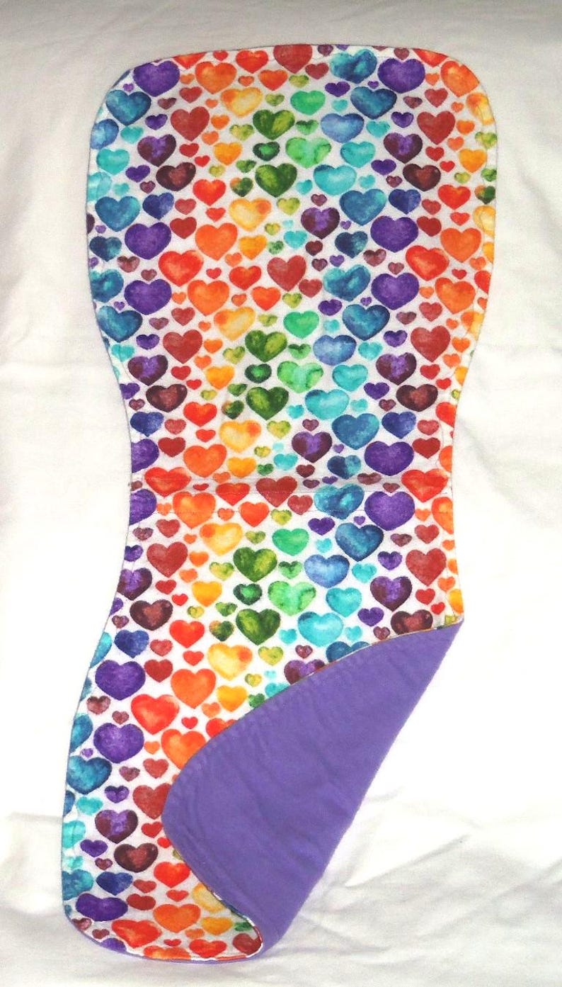May include: A baby burp cloth with a colorful heart pattern in rainbow colors. The hearts are arranged on a white background. The reverse side is a solid purple color. The cloth is made of soft fabric.