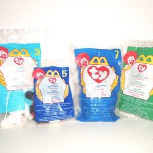 May include: Four Ty plush toys sealed in individual clear plastic McDonald's promotional bags. The bags are labeled with numbers 3, 5, 7, and 11, featuring the McDonald's logo, the Ty heart-shaped logo, and text in English, French, and Spanish.