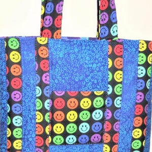 Puede incluir: Un bolso tote con un dise&ntilde;o de patchwork de tela negra con estampado de caras sonrientes coloridas y tela azul con un patr&oacute;n de estampado animal texturizado. El bolso incluye dos asas de tela a juego y un bolsillo frontal cuadrado con caras.