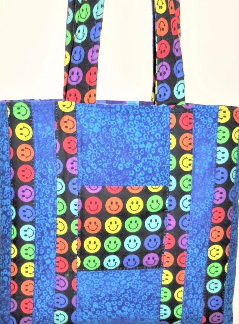 Puede incluir: Un bolso tote con un dise&ntilde;o de patchwork de tela negra con estampado de caras sonrientes coloridas y tela azul con un patr&oacute;n de estampado animal texturizado. El bolso incluye dos asas de tela a juego y un bolsillo frontal cuadrado con caras.