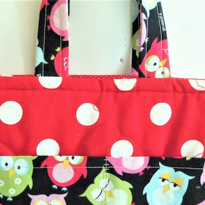 May include: A quilted fabric tote bag featuring a red upper section with white polka dots and a black lower section patterned with colourful owls in shades of pink, green, and blue. The bag includes two matching fabric handles at the top.