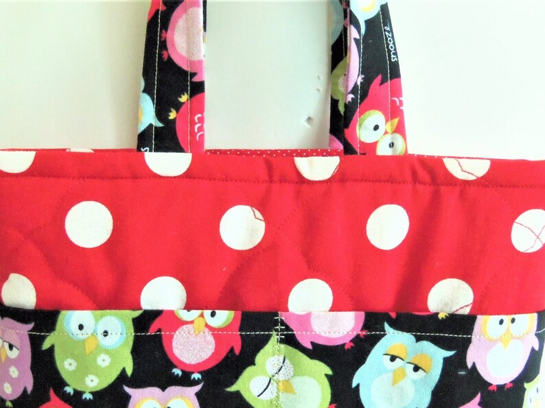 May include: A quilted fabric tote bag featuring a red upper section with white polka dots and a black lower section patterned with colourful owls in shades of pink, green, and blue. The bag includes two matching fabric handles at the top.