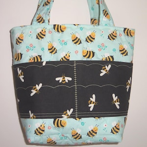 K&ouml;nnte beinhalten: Eine gesteppte Tragetasche mit hellblauem Hintergrund und einem sich wiederholenden Muster aus gelb-schwarzen Bienen und kleinen Blumen. Ein horizontales schwarzes Stoffband mit gewelltem Rand und Bienenmuster ist mittig aufgen&auml;ht.