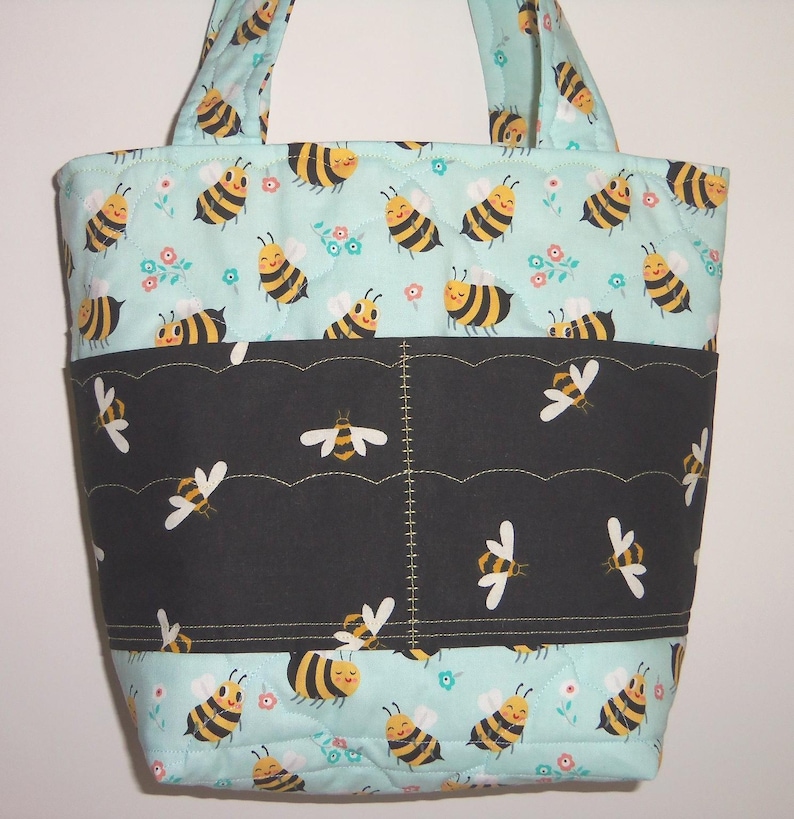 K&ouml;nnte beinhalten: Eine gesteppte Tragetasche mit hellblauem Hintergrund und einem sich wiederholenden Muster aus gelb-schwarzen Bienen und kleinen Blumen. Ein horizontales schwarzes Stoffband mit gewelltem Rand und Bienenmuster ist mittig aufgen&auml;ht.