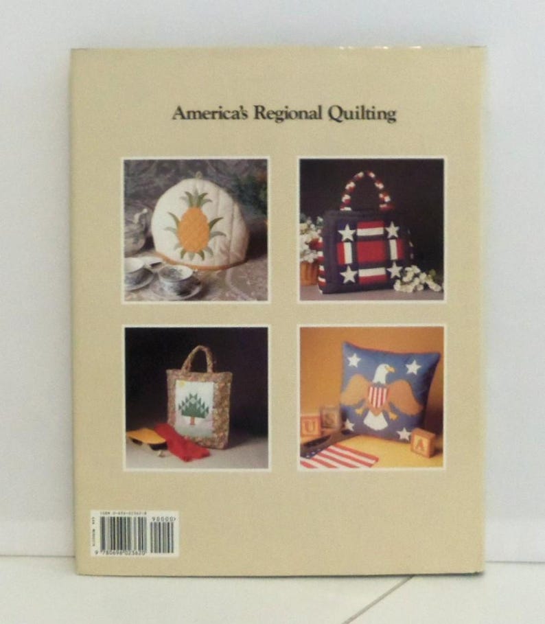 May include: A beige book titled America's Regional Quilting featuring four square photographs on the back cover. The images display a pineapple tea cozy, a red, white, and blue star-patterned tote bag, a tree-motif tote, and an eagle pillow.