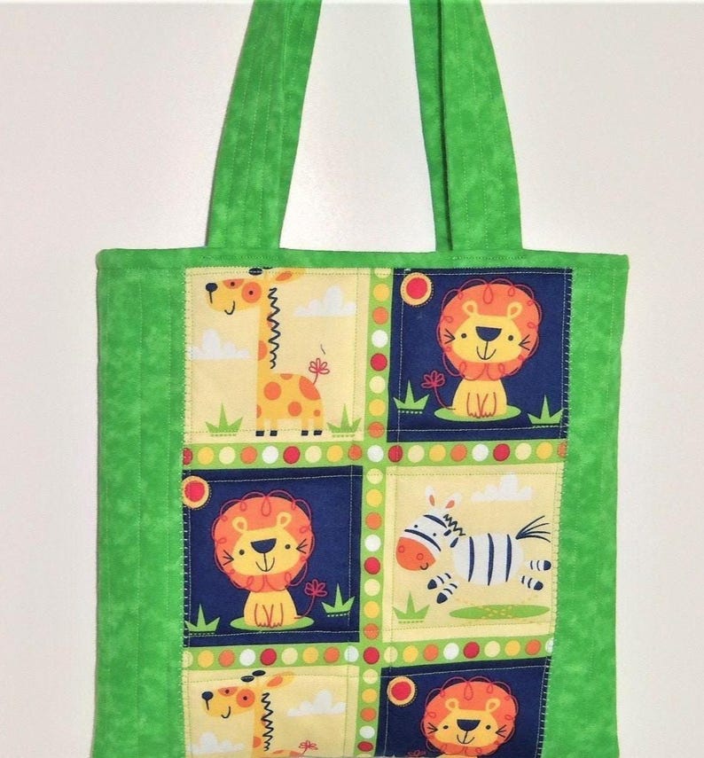May include: A green fabric tote bag featuring a central panel with a grid of squares. The squares display cartoon illustrations of giraffes, lions, and zebras on yellow and dark blue backgrounds, accented by a colorful polka dot border.