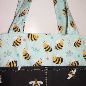 K&ouml;nnte beinhalten: Eine gesteppte Tragetasche mit hellminzgr&uuml;nem Hintergrund und einem sich wiederholenden Muster aus gelb-schwarzen Cartoon-Bienen sowie kleinen rosa und blauen Blumen. Der untere Bereich ist schwarz und mit wei&szlig;en und gelben Bienen bedruckt.