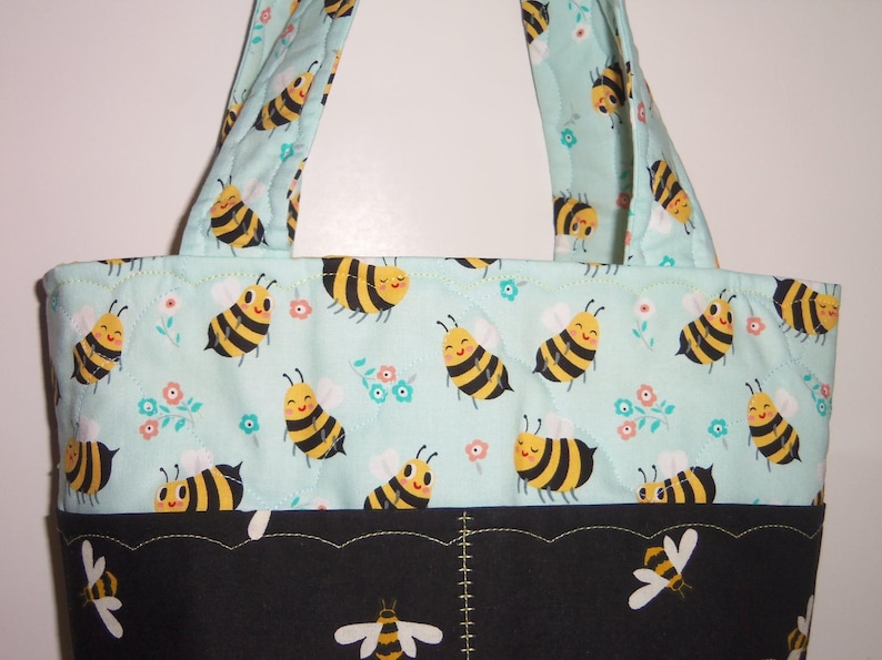 K&ouml;nnte beinhalten: Eine gesteppte Tragetasche mit hellminzgr&uuml;nem Hintergrund und einem sich wiederholenden Muster aus gelb-schwarzen Cartoon-Bienen sowie kleinen rosa und blauen Blumen. Der untere Bereich ist schwarz und mit wei&szlig;en und gelben Bienen bedruckt.