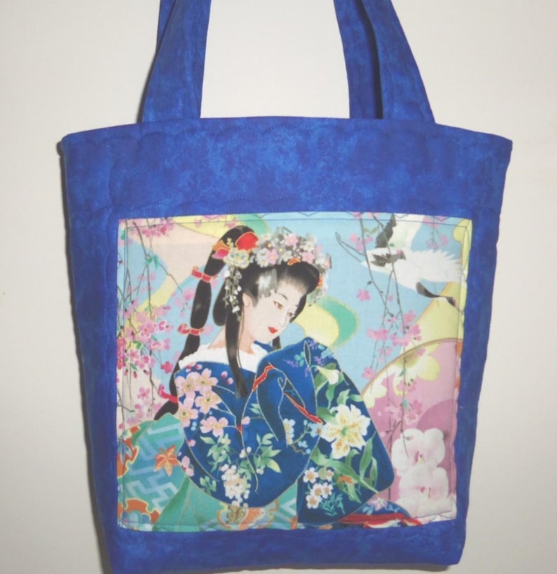 May include: A quilted blue tote bag featuring a central fabric panel with a printed illustration of a person in a floral kimono and hair ornaments, surrounded by cherry blossoms and a crane against a light blue background with two shoulder straps.