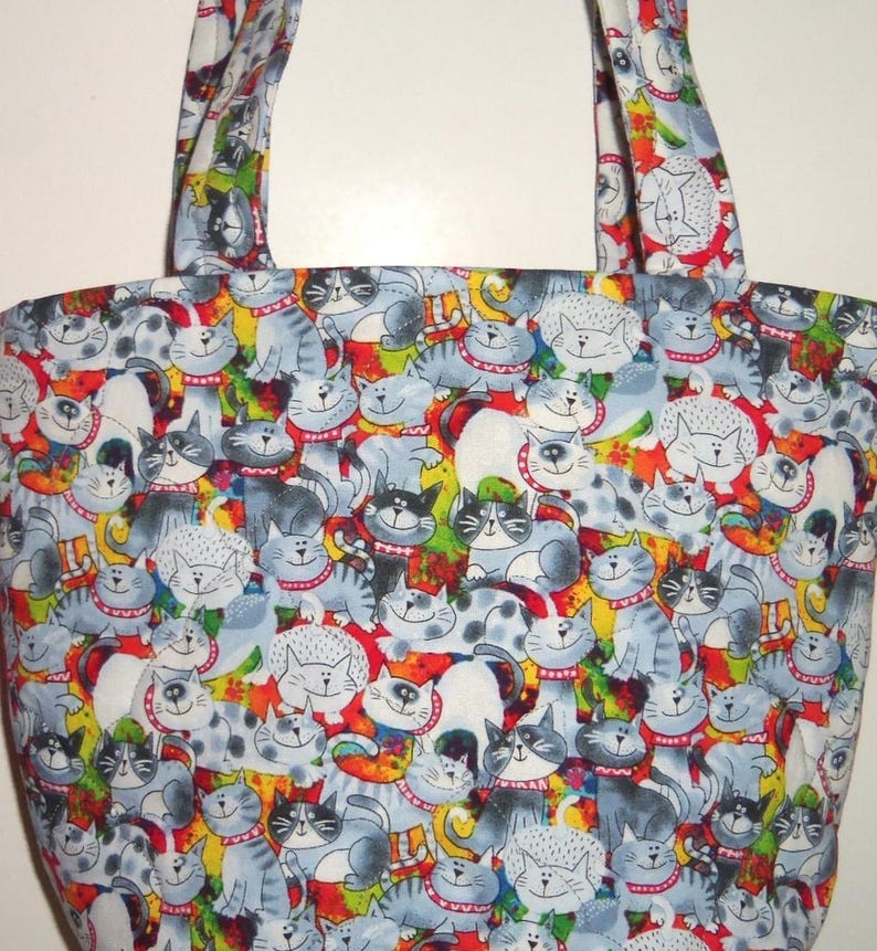 May include: A fabric tote bag featuring a repeating pattern of cartoon cats in shades of gray, white, and black. Each cat wears a red collar with white dots. The background is a vibrant mix of red, yellow, green, and blue abstract shapes.