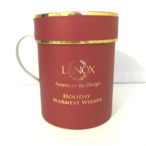 May include: A cylindrical red gift box with a gold rim and a white handle. The front features gold text that reads LENOX American by Design and HOLIDAY WARMEST WISHES. The container has a smooth matte finish and a structured lid design.