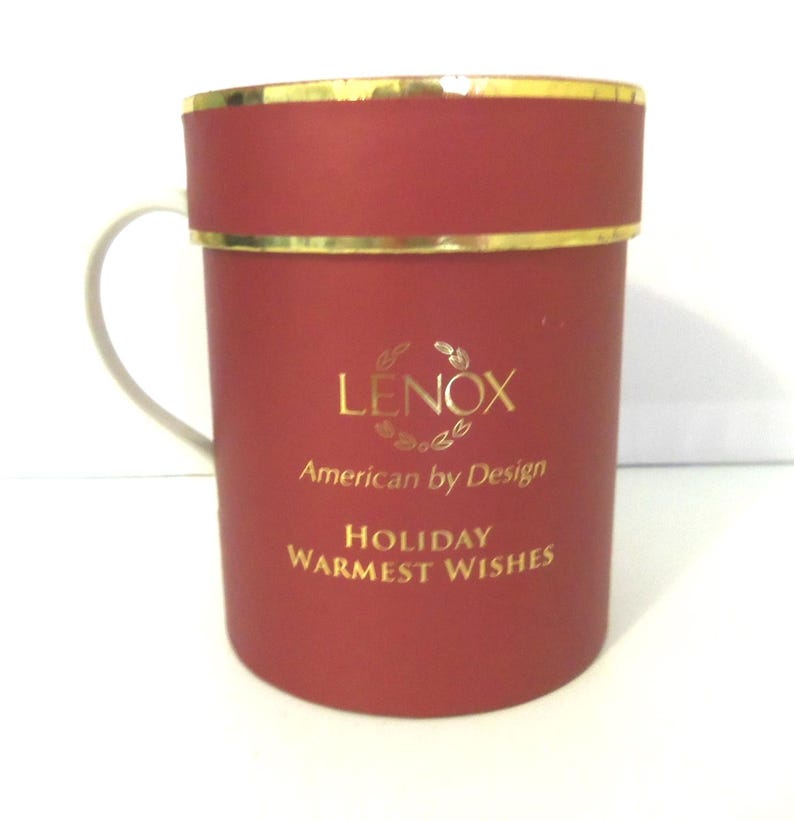 May include: A cylindrical red gift box with a gold rim and a white handle. The front features gold text that reads LENOX American by Design and HOLIDAY WARMEST WISHES. The container has a smooth matte finish and a structured lid design.