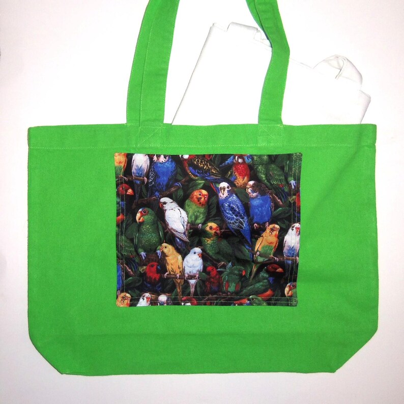 May include: A bright green tote bag featuring a rectangular central panel with a dense, colorful print of various parakeets and parrots perched among dark green foliage. The bag has two matching green fabric shoulder straps.