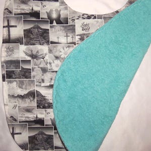 May include: A bib featuring a collage of black and white religious-themed images, including crosses, lighthouses, and flowers, with text such as Light of the World and Walk by Faith Not by Sight. The reverse side is a solid, textured turquoise fabric.