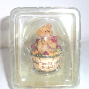 Op de afbeelding: Een bruin teddybeerbeeldje met een rode strik, in een houten mand gevuld met rode appels en ander fruit. De mand heeft de tekst "Thanks a Bushel!!" op de voorkant. Het beeldje zit in een doorzichtige plastic container.