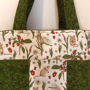 May include: A quilted tote bag featuring a white fabric panel with a repeating pattern of brown mice, red strawberries, and white flowers, contrasted by solid dark green fabric panels and matching dark green shoulder straps.